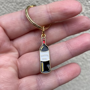 May include: A gold-coloured keyring with a wine bottle charm. The charm is black with a white label that reads "LAFITE 1982". The bottle's top is red, and the keyring is held by a hand.