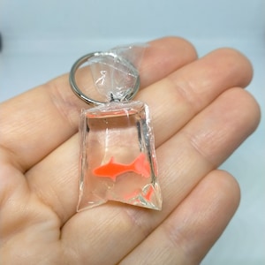 May include: A clear keychain charm resembling a small plastic bag, with a bright orange fish inside. The fish is in a clear liquid. The keychain has a silver-coloured ring.