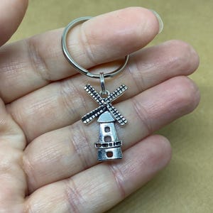 May include: A silver-colored windmill keychain. The windmill charm features a detailed design with blades and a tower. The keychain includes a silver ring for attaching keys. The background is a neutral tan color.