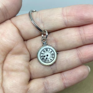 May include: A silver key ring with a round clock charm. The clock face is white with black Roman numeral hour markers and black hands.