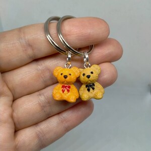 May include: Two small teddy bear keychains with silver key rings. The bears are yellow and have red and black bows.