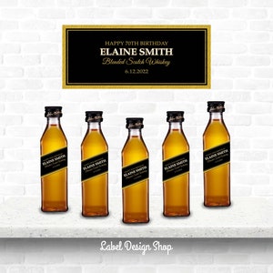 May include: Five miniature bottles of blended Scotch whiskey with black and gold labels, arranged on a white shelf. A sign above reads "Happy 70th Birthday Elaine Smith Blended Scotch Whiskey 6.12.2022."
