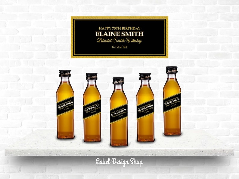 May include: Five miniature bottles of blended Scotch whiskey with black and gold labels, arranged on a white shelf. A sign above reads "Happy 70th Birthday Elaine Smith Blended Scotch Whiskey 6.12.2022."