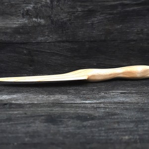 May include: A wooden knife with a light brown handle and a slightly curved blade. The knife is lying on a dark wooden surface.
