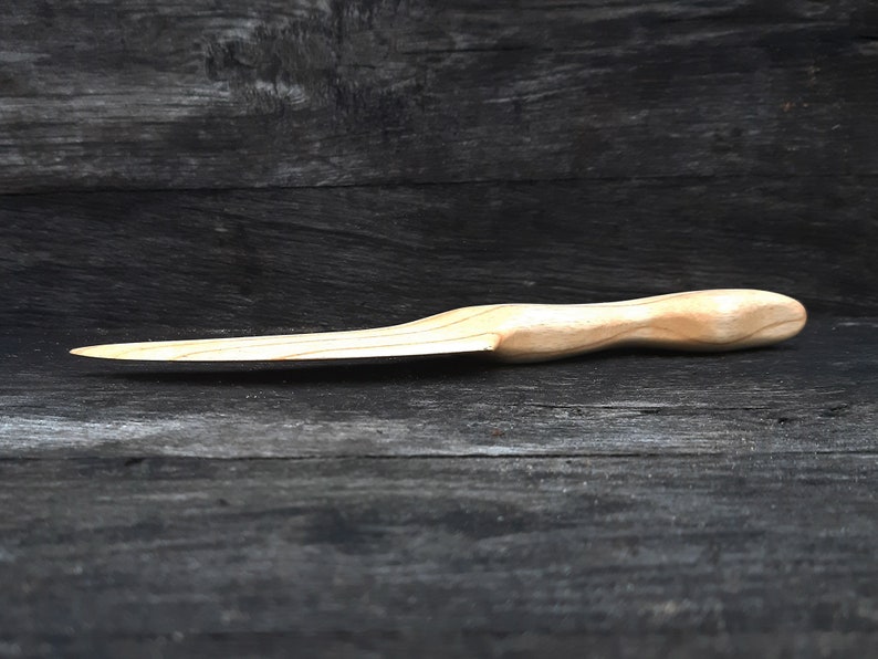 May include: A wooden knife with a light brown handle and a slightly curved blade. The knife is lying on a dark wooden surface.