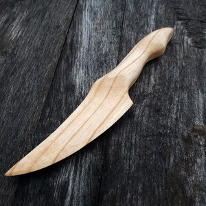 May include: A wooden knife with a light brown finish. The knife has a curved blade and a handle that is slightly angled. The knife is lying on a dark brown wooden surface.