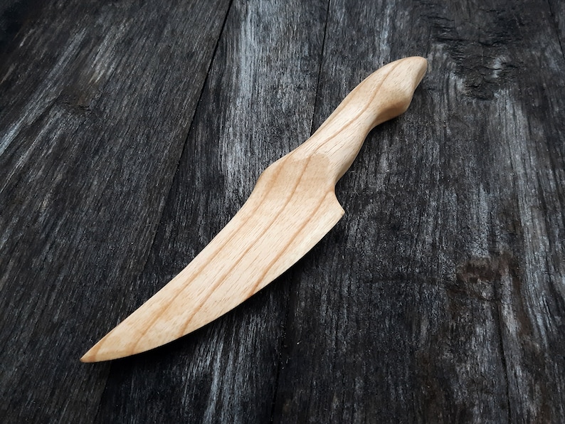 May include: A wooden knife with a light brown finish. The knife has a curved blade and a handle that is slightly angled. The knife is lying on a dark brown wooden surface.