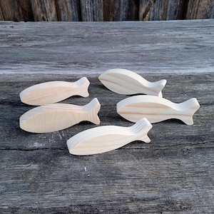 May include: Five wooden fish figurines in various poses. The fish are light-colored, with visible wood grain, and have a simple, stylized design. They are arranged on a weathered, gray wooden surface, suggesting a rustic or nautical theme.