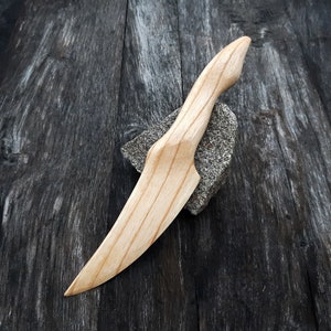 May include: A wooden knife with a natural finish. The knife has a long, thin blade and a rounded handle. The knife is lying on a dark wooden surface.
