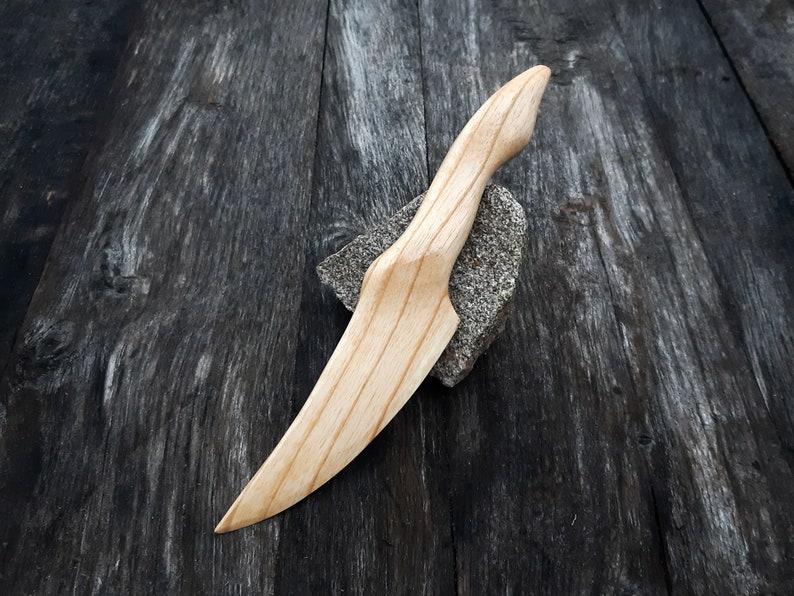May include: A wooden knife with a natural finish. The knife has a long, thin blade and a rounded handle. The knife is lying on a dark wooden surface.