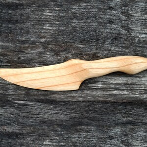 May include: A wooden butter knife with a curved handle and a pointed blade. The knife is made of light wood and has a natural finish.