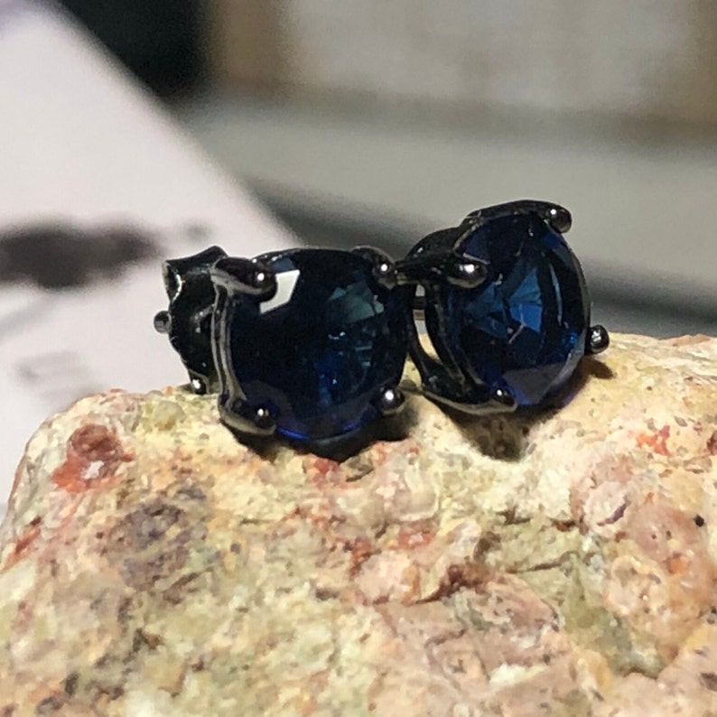 May include: A pair of stud earrings featuring round, faceted dark blue stones set in black metal prongs. The earrings are positioned side by side, showcasing the deep blue color of the stones and the dark, metallic finish of the setting.