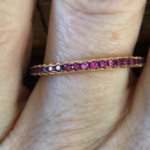 May include: A gold ring featuring a continuous line of small, bright pink gemstones. The ring's band has a textured, rope-like pattern. The gemstones are set closely together, creating a sparkling effect. The ring is shown on a person's finger.