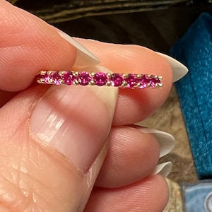 Op de afbeelding: Een gouden bandring met een doorlopende rij kleine, ronde, helder roze edelstenen. De ring wordt tussen de vingers gehouden, waardoor de levendige kleur van de edelstenen en het delicate ontwerp van de ring worden benadrukt.