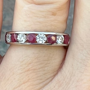 Puede incluir: Un anillo de banda plateado con piedras preciosas rojas y claras alternas. El anillo presenta un patrón continuo de piedras preciosas engarzadas alrededor de la banda. Las piedras están engarzadas en un engaste de canal.