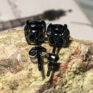 May include: A pair of stud earrings featuring round, deep blue faceted stones set in black metal prongs. The earrings include thin black posts and matching black butterfly-style friction backs, showing the rear view of the jewelry construction.