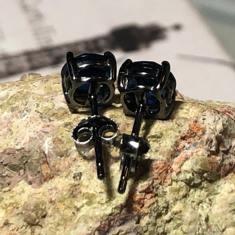 May include: A pair of stud earrings featuring round, deep blue faceted stones set in black metal prongs. The earrings include thin black posts and matching black butterfly-style friction backs, showing the rear view of the jewelry construction.