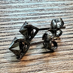 May include: A pair of black diamond-shaped stud earrings. The earrings have a dark, metallic finish and are set on thin posts with butterfly clasps. The earrings are displayed on a light-colored, textured surface.
