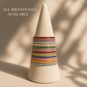 Könnte beinhalten: Ein Stapel aus elf dünnen Metallringen, die jeweils mit einer Reihe kleiner, runder Edelsteine in verschiedenen Farben besetzt sind, präsentiert auf einem beigen Kegel. Der Text ALL BIRTHSTONES AVAILABLE steht oben links.