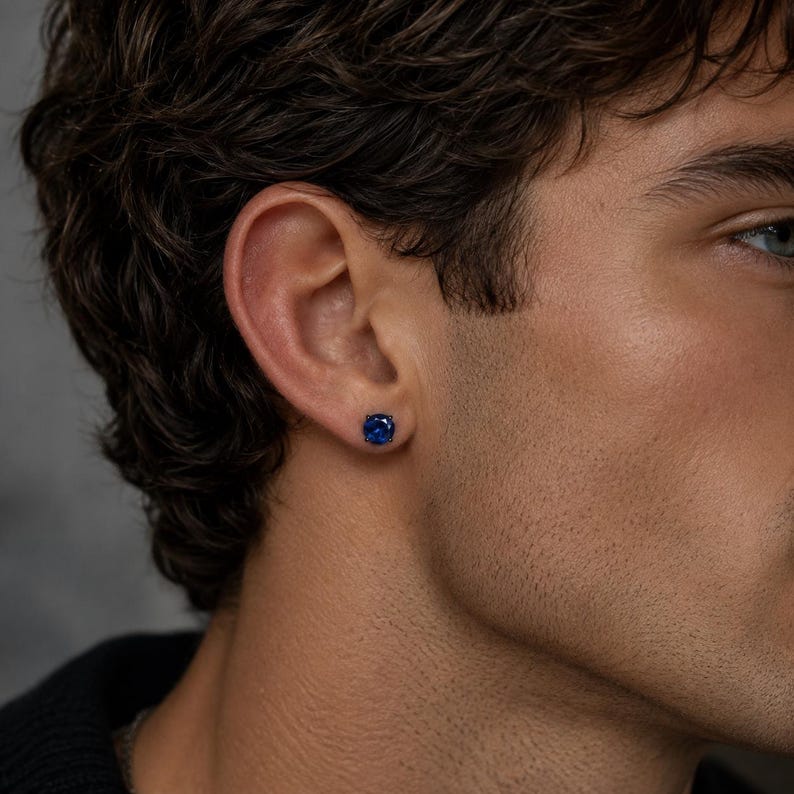 May include: A round, deep blue stud earring featuring a faceted gemstone set in a dark metal prong setting. The earring is positioned in the earlobe, showcasing a brilliant, saturated blue color and a circular cut that reflects light.