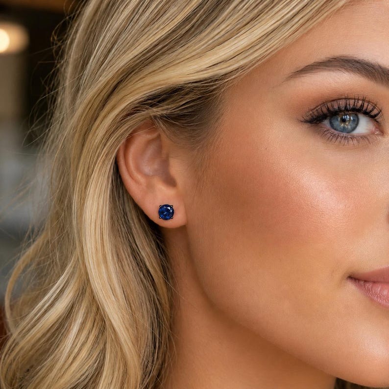 May include: A small, round, deep blue stud earring featuring a faceted gemstone set in a dark metal prong setting. The earring is positioned centrally on the earlobe, showcasing a vibrant, saturated blue color and a circular, polished cut.