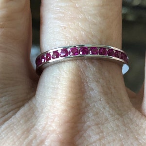 Puede incluir: Un anillo de plata con una banda de pequeñas y vibrantes piedras preciosas rojas. El anillo está engastado con múltiples rubíes de talla redonda, creando una fila continua de color. El anillo se muestra en un dedo.