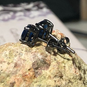 May include: A pair of stud earrings featuring round, deep blue faceted stones set in dark, gunmetal-toned metal prongs. The earrings include matching dark metal posts and butterfly-style friction backs, showcasing a clear, polished gemstone finish.