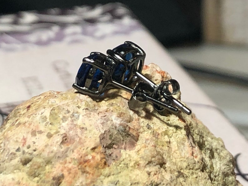 May include: A pair of stud earrings featuring round, deep blue faceted stones set in dark, gunmetal-toned metal prongs. The earrings include matching dark metal posts and butterfly-style friction backs, showcasing a clear, polished gemstone finish.