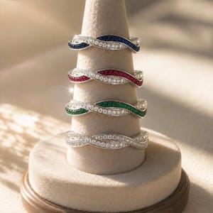 May include: Four silver-toned twisted bands stacked vertically on a conical display. Each ring features a dual-strand design with one row of clear stones and one row of colored stones, including blue, red, green, and a final band of all clear stones.