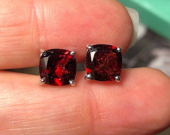 May include: A pair of silver stud earrings with red gemstone accents. The gemstones are square-shaped and have a faceted cut.