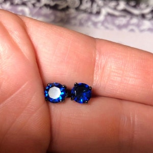 May include: A pair of round-cut stud earrings featuring vibrant blue stones set in dark, black-toned metal prongs. The circular blue gems are faceted to reflect light, creating a deep, saturated color contrast against the dark metal settings.