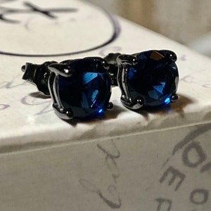 May include: A pair of stud earrings featuring round, faceted dark blue stones set in dark metal prongs. The earrings are positioned side by side, showcasing the deep blue color and the reflective, multi-faceted surface of the circular gemstones.