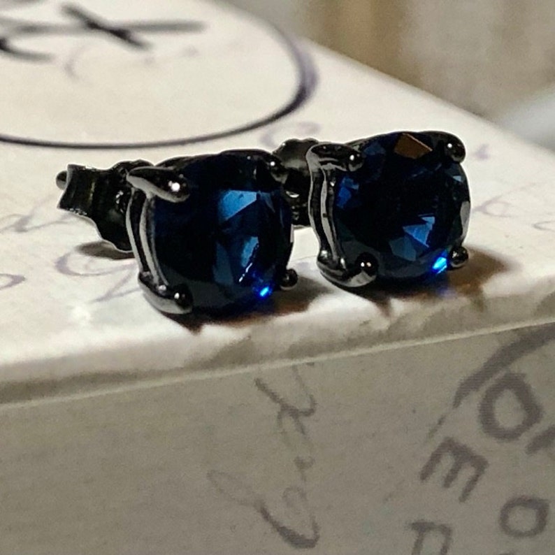 May include: A pair of stud earrings featuring round, faceted dark blue stones set in dark metal prongs. The earrings are positioned side by side, showcasing the deep blue color and the reflective, multi-faceted surface of the circular gemstones.