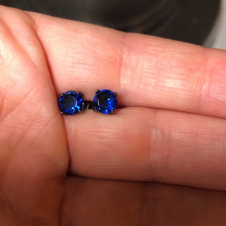 May include: A pair of stud earrings featuring round, faceted, deep blue stones set in dark, black-toned metal prongs. The earrings are small, circular, and display a vibrant, reflective blue color against the dark metal setting.