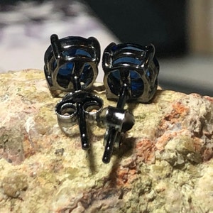 May include: A pair of stud earrings featuring round, deep blue faceted stones set in dark, gunmetal-toned metal prongs. The earrings include matching dark metal posts and butterfly-style friction backs, showing the rear construction of the jewelry.
