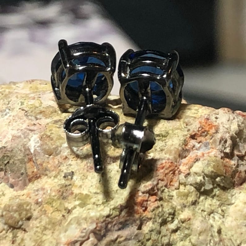 May include: A pair of stud earrings featuring round, deep blue faceted stones set in dark, gunmetal-toned metal prongs. The earrings include matching dark metal posts and butterfly-style friction backs, showing the rear construction of the jewelry.