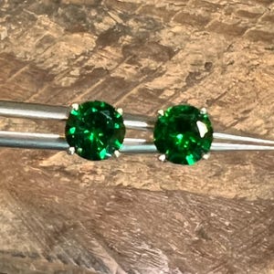 Può includere: Un paio di orecchini a bottone rotondi verde smeraldo. Ogni orecchino presenta una gemma verde vibrante incastonata in una montatura in metallo argentato o bianco. Gli orecchini sono tenuti da una pinzetta su uno sfondo di legno.