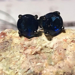 May include: A pair of dark blue stud earrings with a faceted, round gemstone design. The earrings are set in a dark metal with four small prongs holding the gemstone in place. The earrings are displayed on a textured, light brown surface.