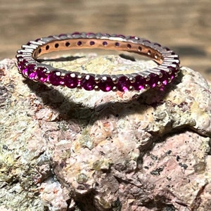 May include: A silver eternity ring with a band of vibrant, round, magenta-colored gemstones. The ring is set against a textured, light brown and gray stone surface, creating a contrast of textures and colors. The ring's design is simple and elegant.