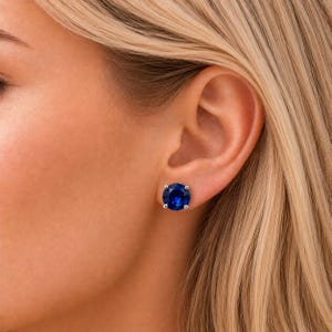 May include: A close-up of a person's ear with a large, round, deep blue stud earring. The earring is set in a silver or white metal. The person has blonde hair and fair skin.