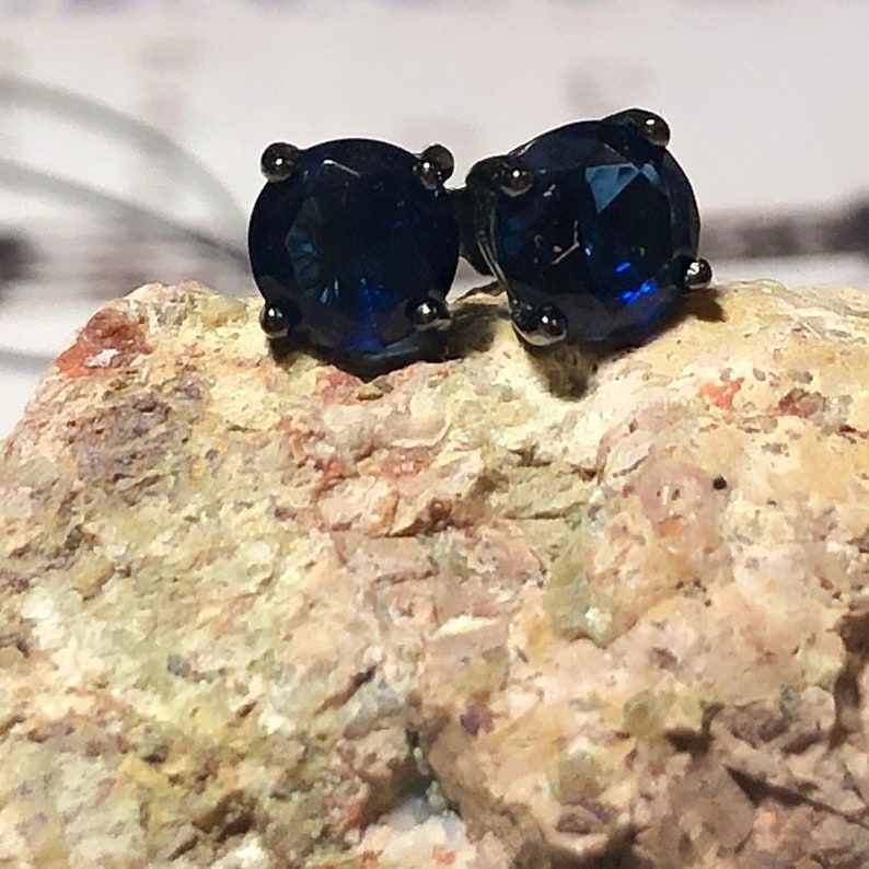 May include: A pair of round-cut dark blue stud earrings set in black metal prongs. The earrings are positioned on a textured, light-colored, porous rock surface with visible mineral flecks and uneven edges, highlighting the deep blue gemstone color.