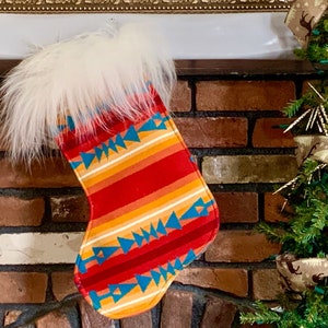 Puede incluir: Un calcetín navideño con un colorido diseño del suroeste. El calcetín presenta un patrón de rayas rojas, naranjas y amarillas con detalles de triángulos azules. La parte superior del calcetín está adornada con suave piel sintética blanca.