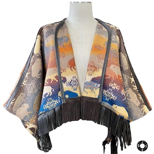 May include: A brown, blue and orange patterned poncho with fringe trim. The poncho is made of a soft, woven fabric and features a geometric design with images of buffalo.