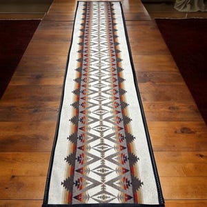 Puede incluir: Camino de mesa rectangular con un patrón geométrico en tonos blancos, marrones, rojos y grises. El camino de mesa tiene un borde negro y está colocado sobre una mesa de madera. El diseño presenta formas repetidas de diamantes y triángulos.