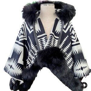 May include: A black and white patterned poncho with a hood and fur trim. The poncho features a geometric design and is trimmed with black fur around the hood and front edges. The garment is displayed on a mannequin.