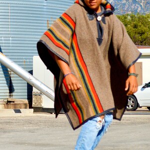May include: A young person wearing a brown and red striped poncho with blue jeans and black sneakers is riding a skateboard. The person is standing on a paved surface in front of a large metal silo.
