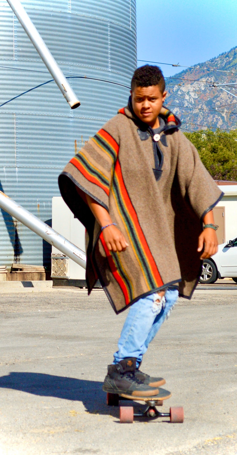 May include: A young person wearing a brown and red striped poncho with blue jeans and black sneakers is riding a skateboard. The person is standing on a paved surface in front of a large metal silo.