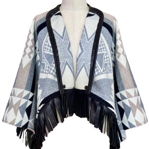 May include: A geometric patterned poncho in shades of gray, black, and white, displayed on a mannequin. The poncho features a black leather trim and fringe along the bottom edge. The design includes diamond and triangle shapes.