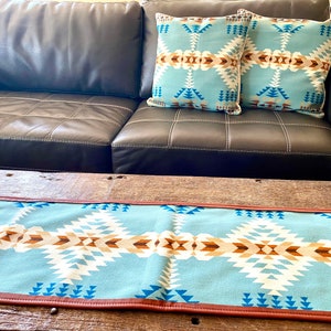 Puede incluir: Un camino de mesa azul y marrón con estampado y ribete de cuero marrón. El camino de mesa está sobre una mesa de madera delante de un sofá de cuero marrón con cojines decorativos azules y marrones.