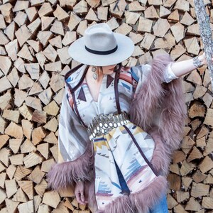 May include: A woman wearing a white hat, a patterned poncho with fur trim, and a silver belt with a large buckle. She is standing in front of a wall of stacked firewood.